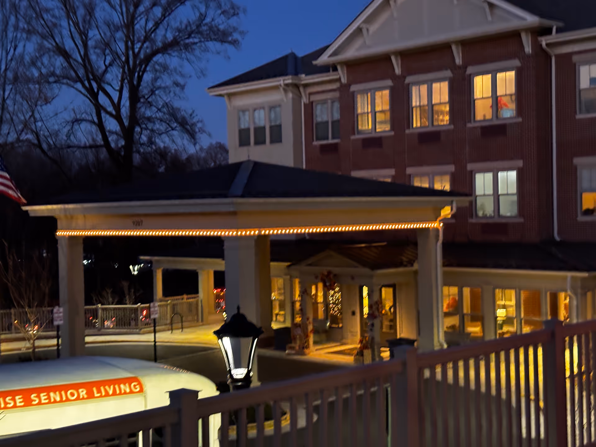 Evening view of the entrance to a senior living facility with a covered drop-off area lit by string lights. The building is multi-story with several windows illuminated from inside. A lamp post and part of a vehicle with 'Sunrise Senior Living' signage are visible in the foreground.