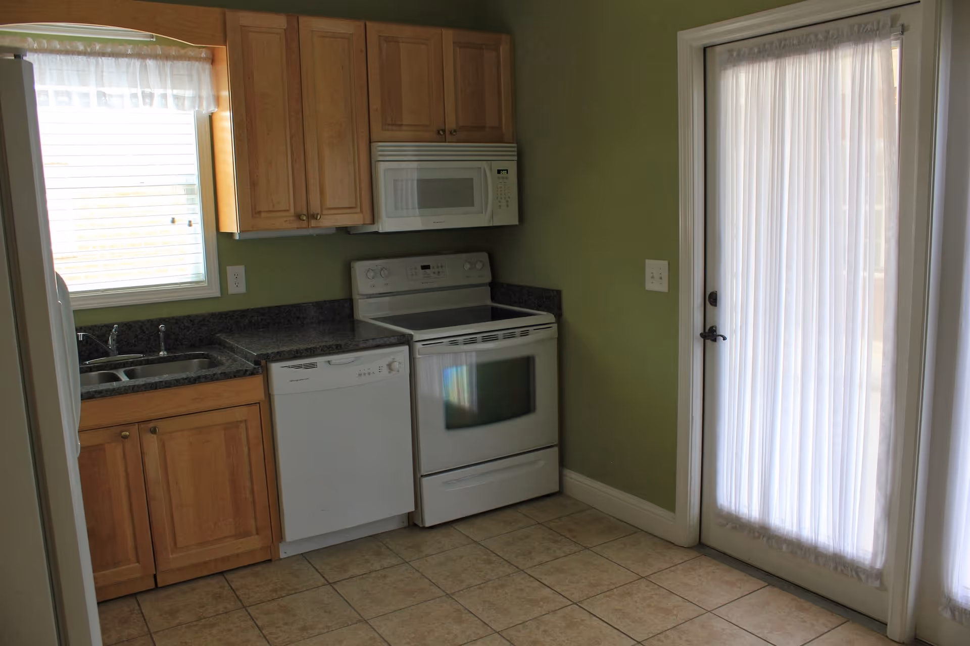 A kitchen with light wood cabinets, a white microwave above a white electric stove, a white dishwasher, a double sink under a window with a sheer curtain, green walls, and a glass door with sheer curtains leading outside. The floor is tiled with beige tiles.