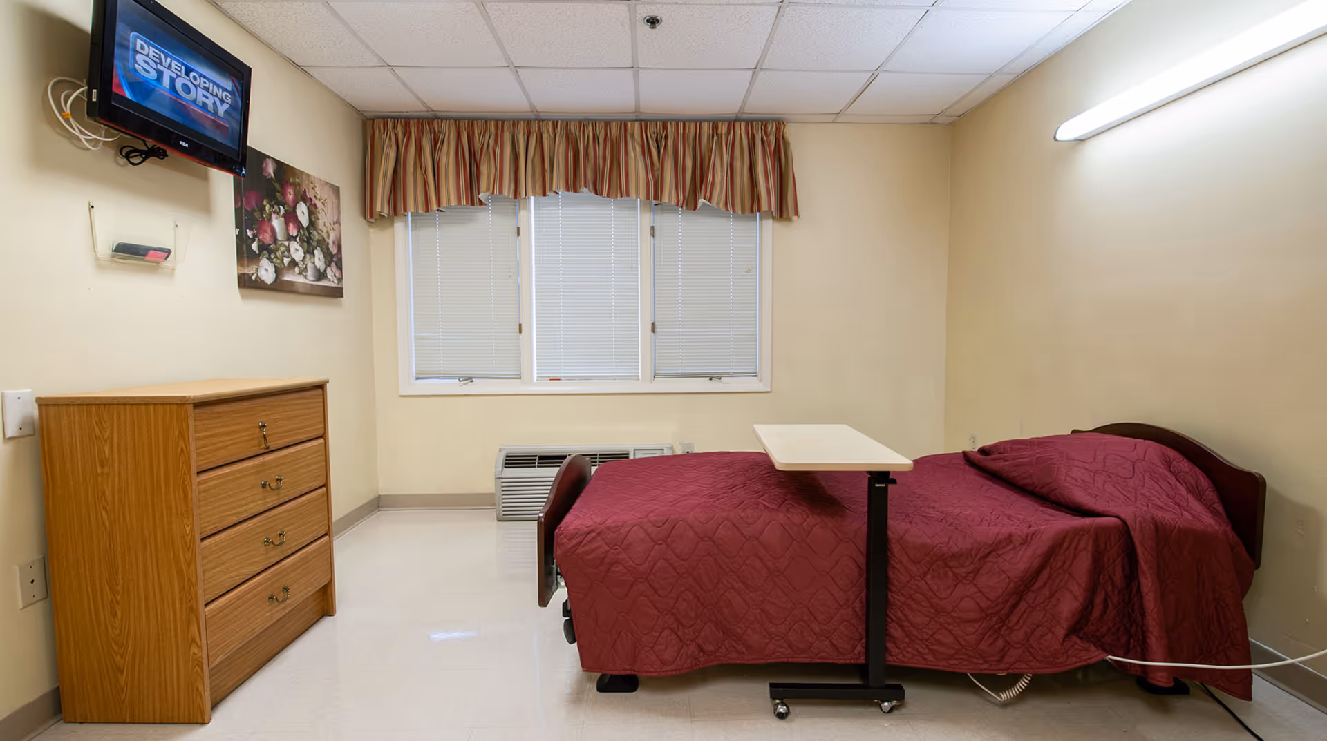 A simple room with a single bed covered in a maroon quilt, a wooden dresser with four drawers, a wall-mounted television displaying the text 'DEVELOPING STORY', a floral painting, and a window with closed blinds and striped valance. There is a rolling overbed table positioned over the bed and a fluorescent light fixture on the wall.