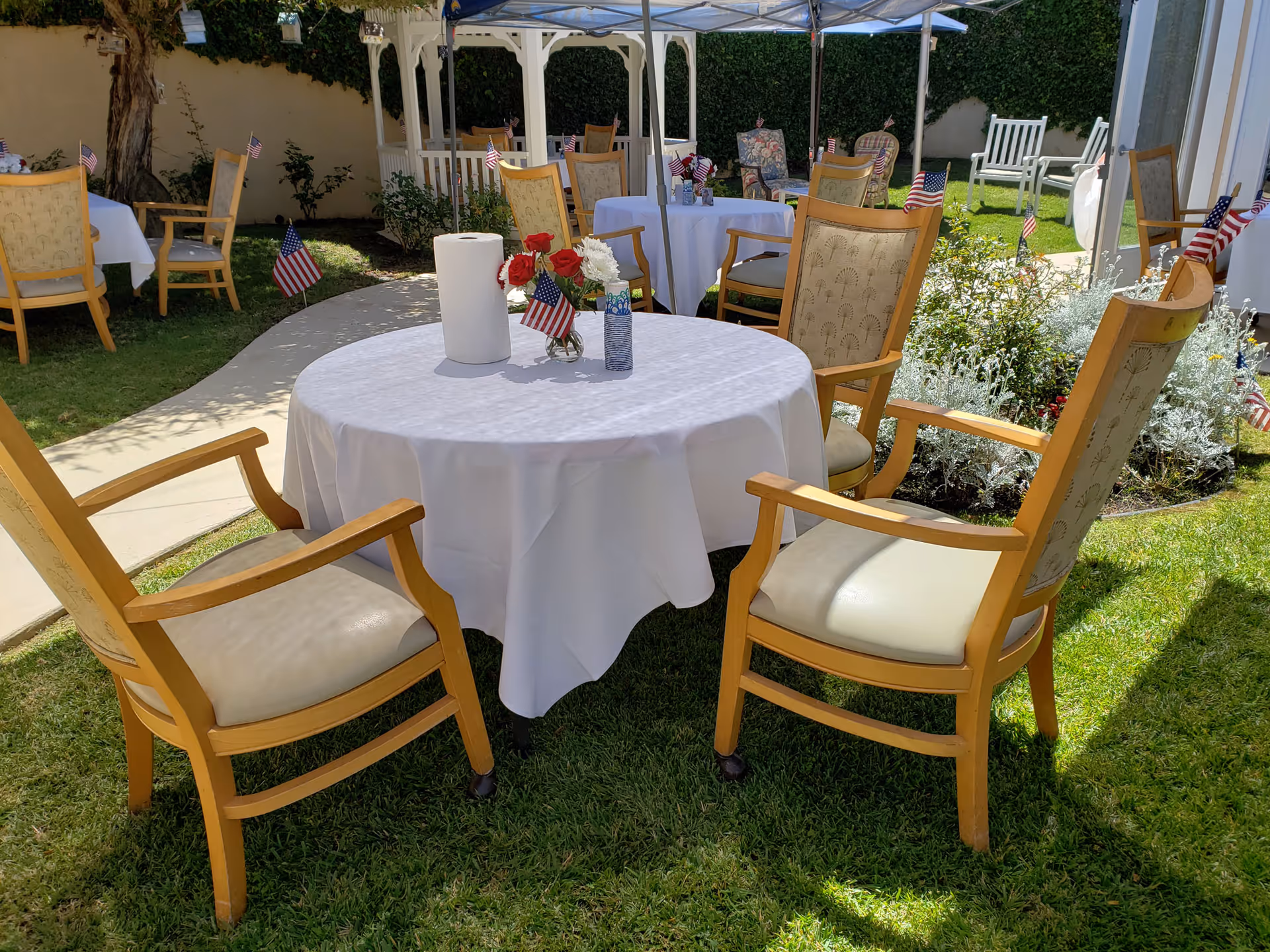 Outdoor seating area with round tables covered in white tablecloths, surrounded by wooden chairs with cushioned seats. Small American flags and flower arrangements are placed on the tables. The area is grassy with a paved walkway and some garden plants in the background, under a blue canopy.