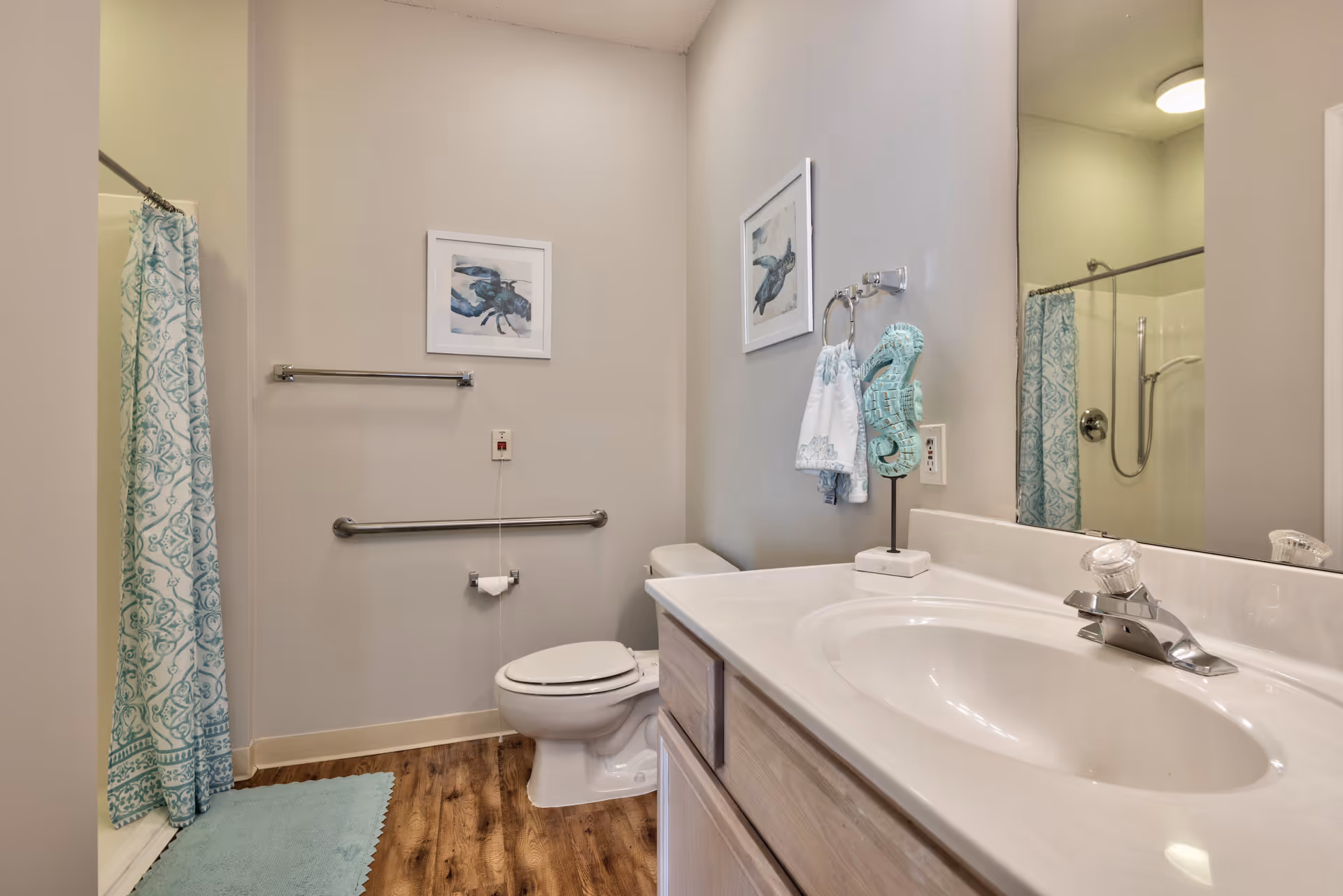 A bathroom with a white toilet, a sink with a countertop, a large mirror, and a shower with a blue and white patterned curtain. The walls are light-colored and decorated with framed artwork. There is a towel hanging on a ring next to the sink and a blue bath mat on the wooden floor.