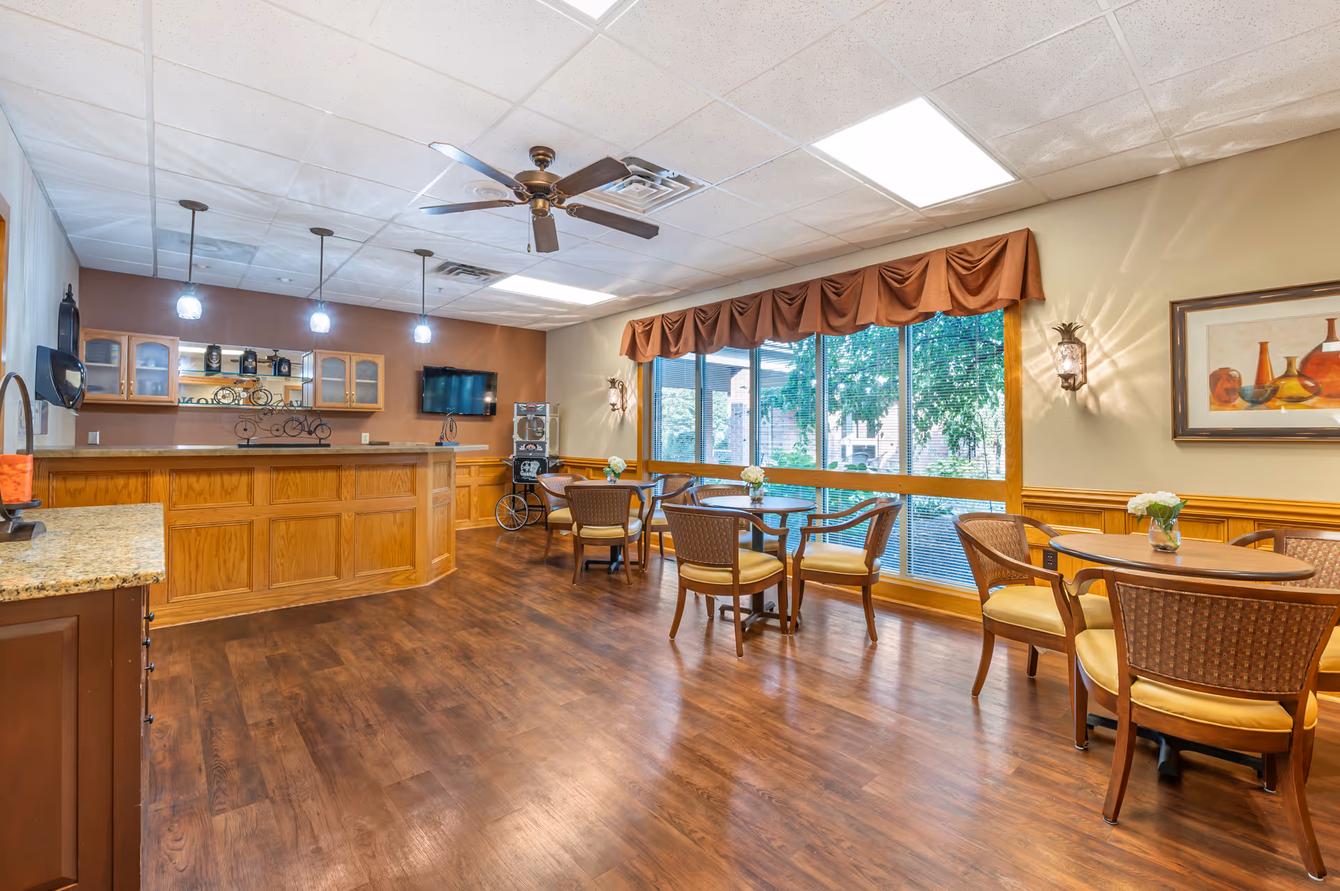 A spacious room with wooden flooring and a ceiling fan, featuring a counter with wooden cabinets and three hanging lights above it. There are several round tables with chairs arranged near large windows with brown valances, allowing natural light to enter. The walls are decorated with a framed painting and wall sconces, and a flat-screen TV is mounted on the far wall.
