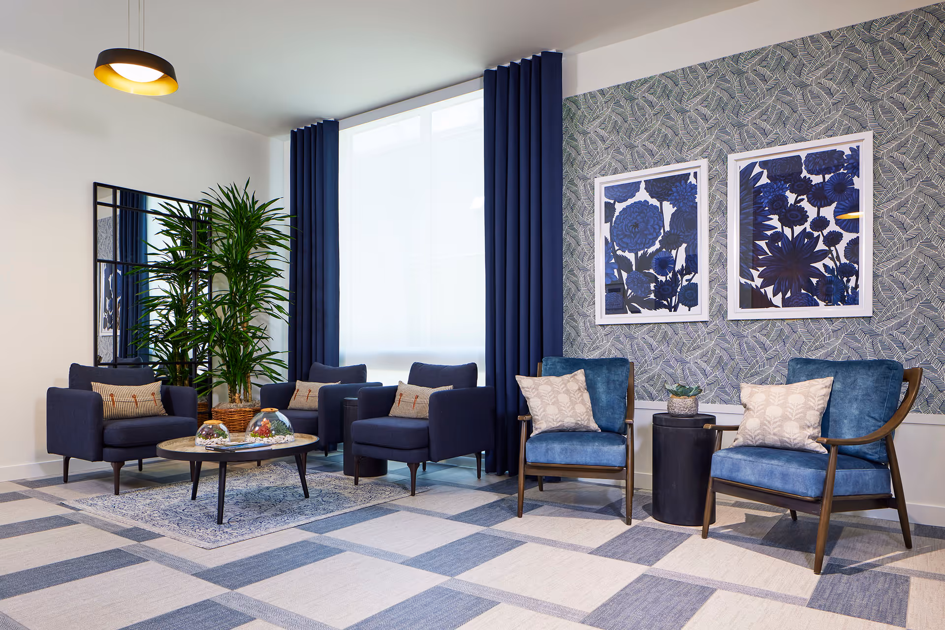 Modern seating area with blue armchairs, a round coffee table, a large potted plant, and framed blue botanical prints on patterned wallpaper.