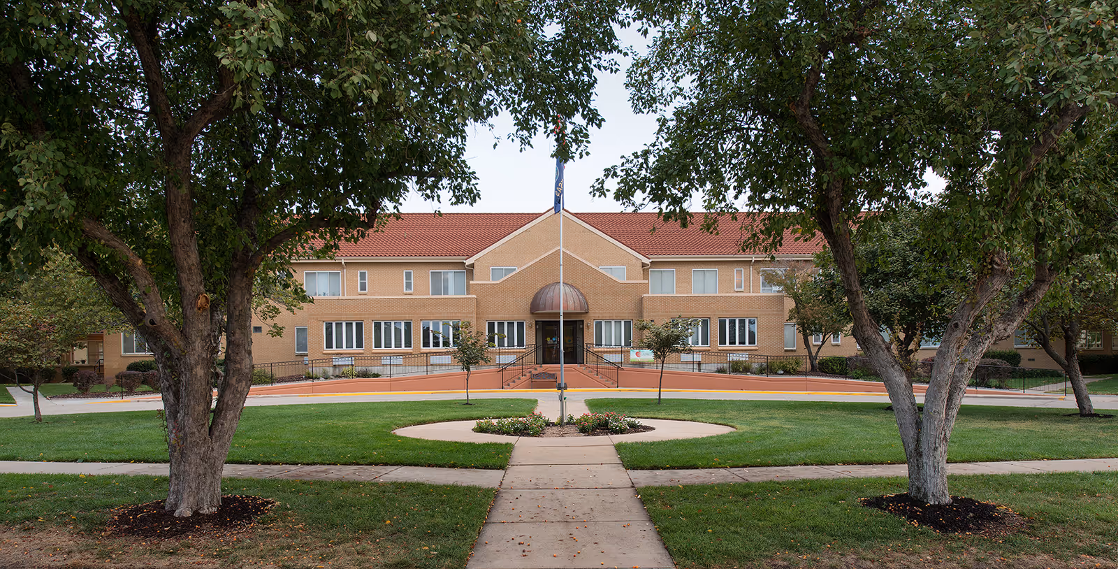 Front exterior view of Newton Presbyterian Manor, a two-story brick building with a red roof, surrounded by green lawns and trees, with a flagpole and a circular flower bed in the center of the walkway leading to the entrance.