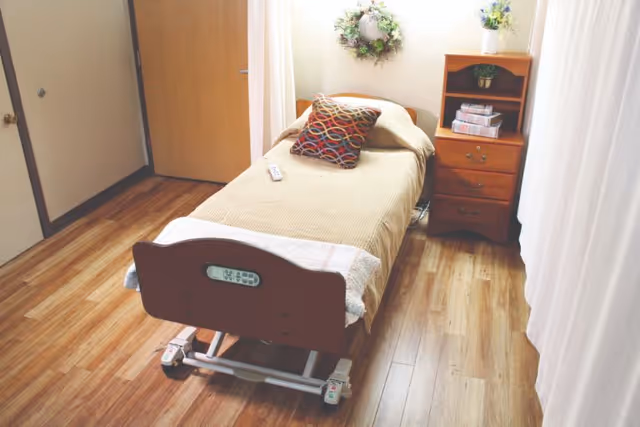 A single adjustable hospital bed with a patterned pillow and remote in a small furnished patient bedroom with a wooden nightstand and wood floors.