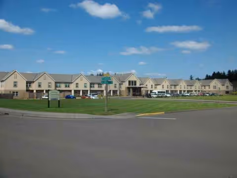 Row of beige senior living buildings with a grassy lawn, parked cars, and blue sky.