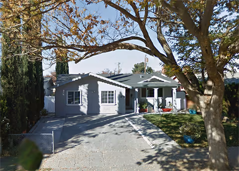 A single-story residential care home exterior with driveway, front lawn, and trees.