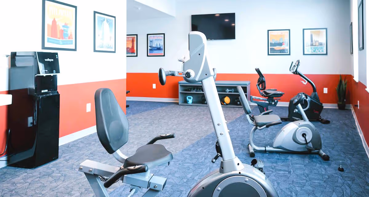 Small fitness room with recumbent exercise bikes, wall-mounted TV, framed art, and a water dispenser.