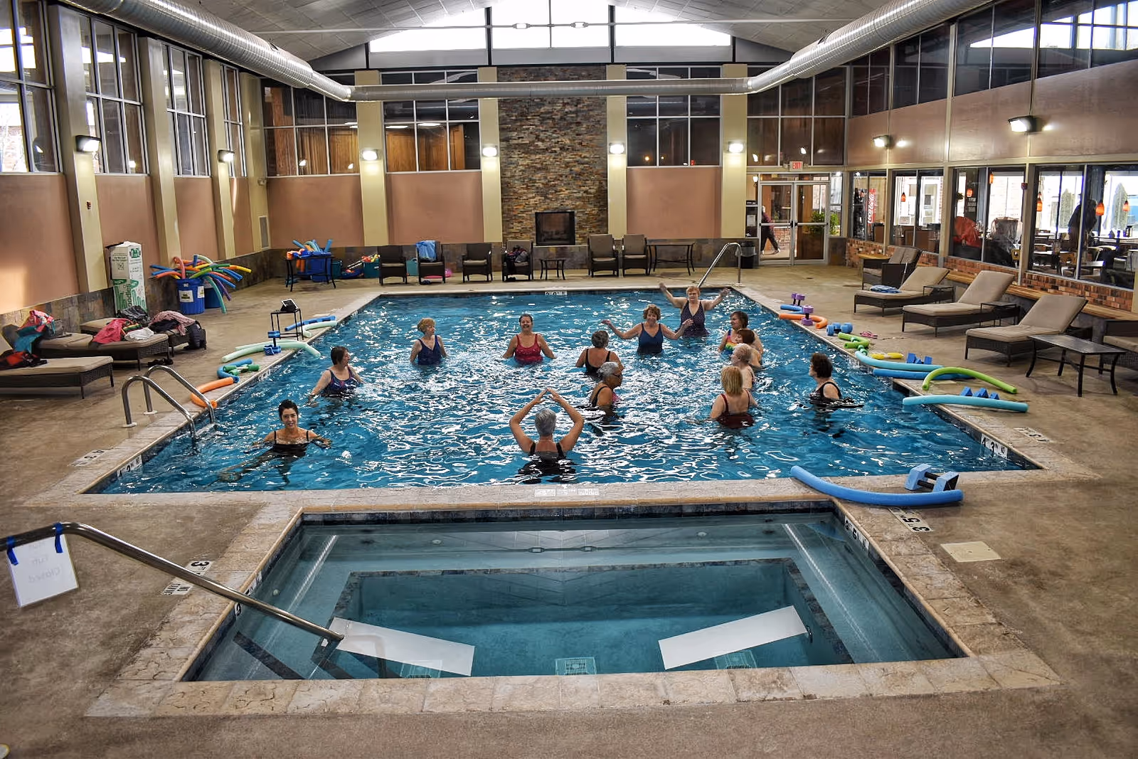 Indoor swimming pool area with a group of people participating in a water exercise class. The pool is surrounded by lounge chairs, pool noodles, and exercise equipment. There is a smaller hot tub in the foreground and large windows along the walls letting in natural light.