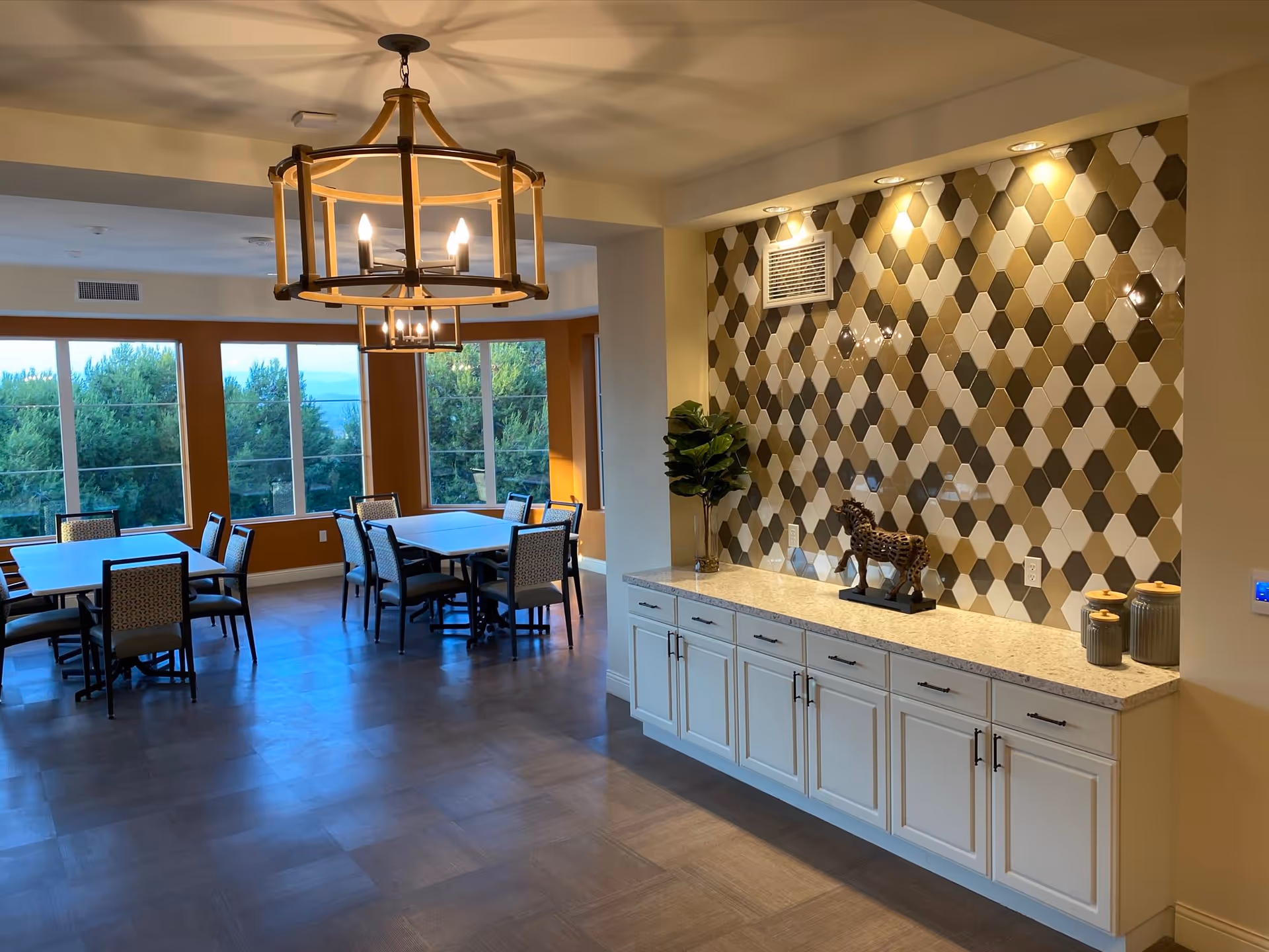 Interior view of a dining area in Silvergate Rancho Bernardo featuring several tables and chairs near large windows with a view of trees outside. The room has a modern chandelier hanging from the ceiling and a decorative wall with hexagonal tiles in shades of beige, brown, and white. Below the tiled wall is a long countertop with white cabinets, a small plant, a horse figurine, and two decorative jars.