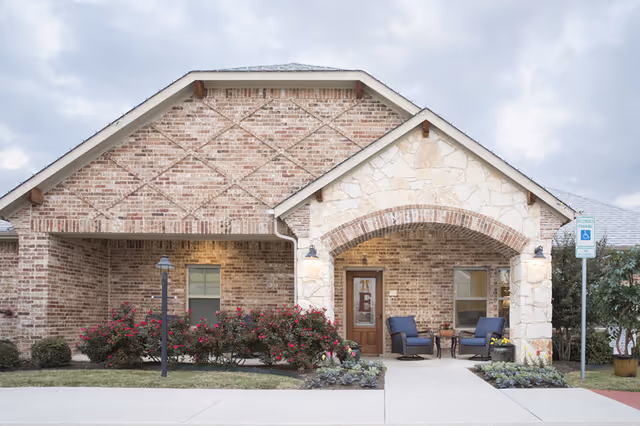 Front exterior view of a single-story brick and stone building with a covered porch featuring two blue cushioned chairs and a small table. There are bushes with pink flowers and a reserved parking sign for handicapped parking near the entrance.
