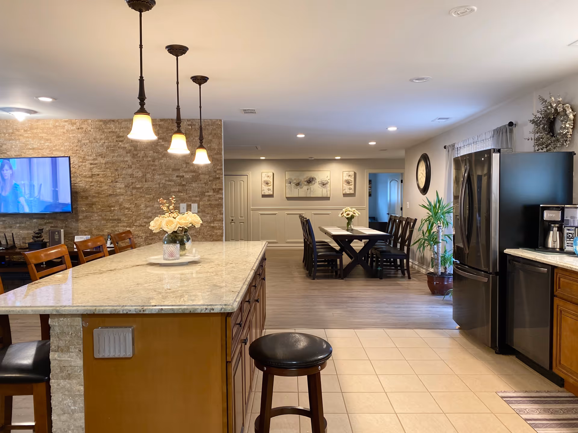 Interior view of a senior living facility showing a kitchen area with a large island countertop, bar stools, pendant lights, and a refrigerator. In the background, there is a dining area with a long table, chairs, wall clock, and decorative wall art. The space is well-lit with recessed lighting and has a mix of tile and wood flooring.
