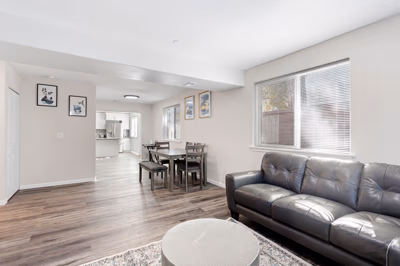 A bright and clean living area featuring a black leather sofa, a round white coffee table on a patterned rug, and a dining table with chairs and a bench. The room has wood flooring, white walls with framed artwork, and large windows with blinds letting in natural light. The kitchen is visible in the background.