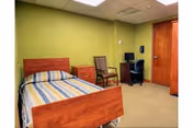 Private bedroom with a single wooden bed with striped bedding, green walls, a dresser, chairs, a desk with a computer, and a closed wooden door.
