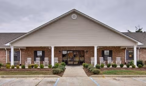 Front exterior view of a single-story brick building with a covered entrance supported by white columns. There are several white rocking chairs lined up on the porch, and small shrubs and plants are arranged along the front of the building. The sky is overcast.