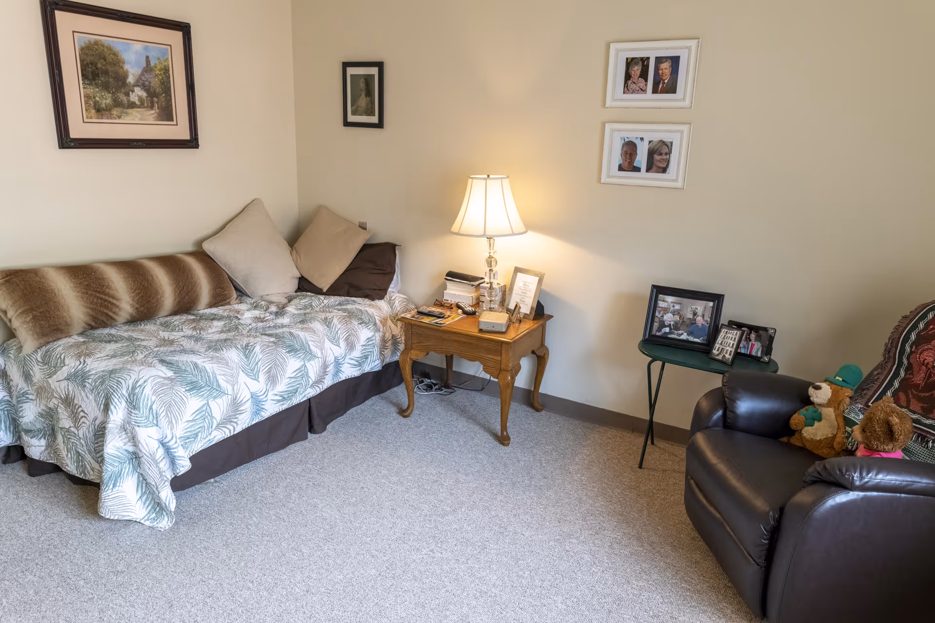 A cozy room with a daybed covered in a leafy patterned blanket and several pillows. Next to the bed is a wooden side table with a lamp, books, and framed photos. On the right side, there is a black leather armchair with stuffed teddy bears and a small green side table holding more framed photos. The walls are decorated with framed pictures and photographs.