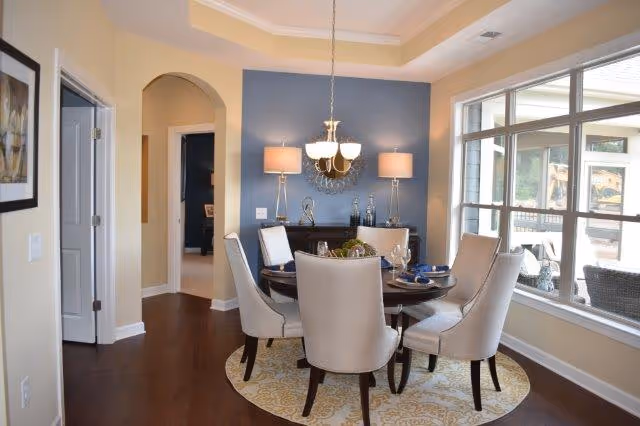 A dining room with a round wooden table surrounded by six beige upholstered chairs on a patterned round rug. The room features a blue accent wall with two table lamps and a decorative mirror. A chandelier hangs above the table, and large windows on the right side let in natural light, showing an outdoor patio area.
