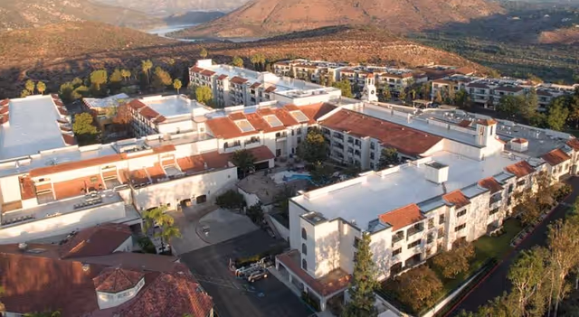 Aerial view of Casa de las Campanas senior living facility showing multiple connected buildings with white roofs and red tile accents, surrounded by trees and mountains in the background.