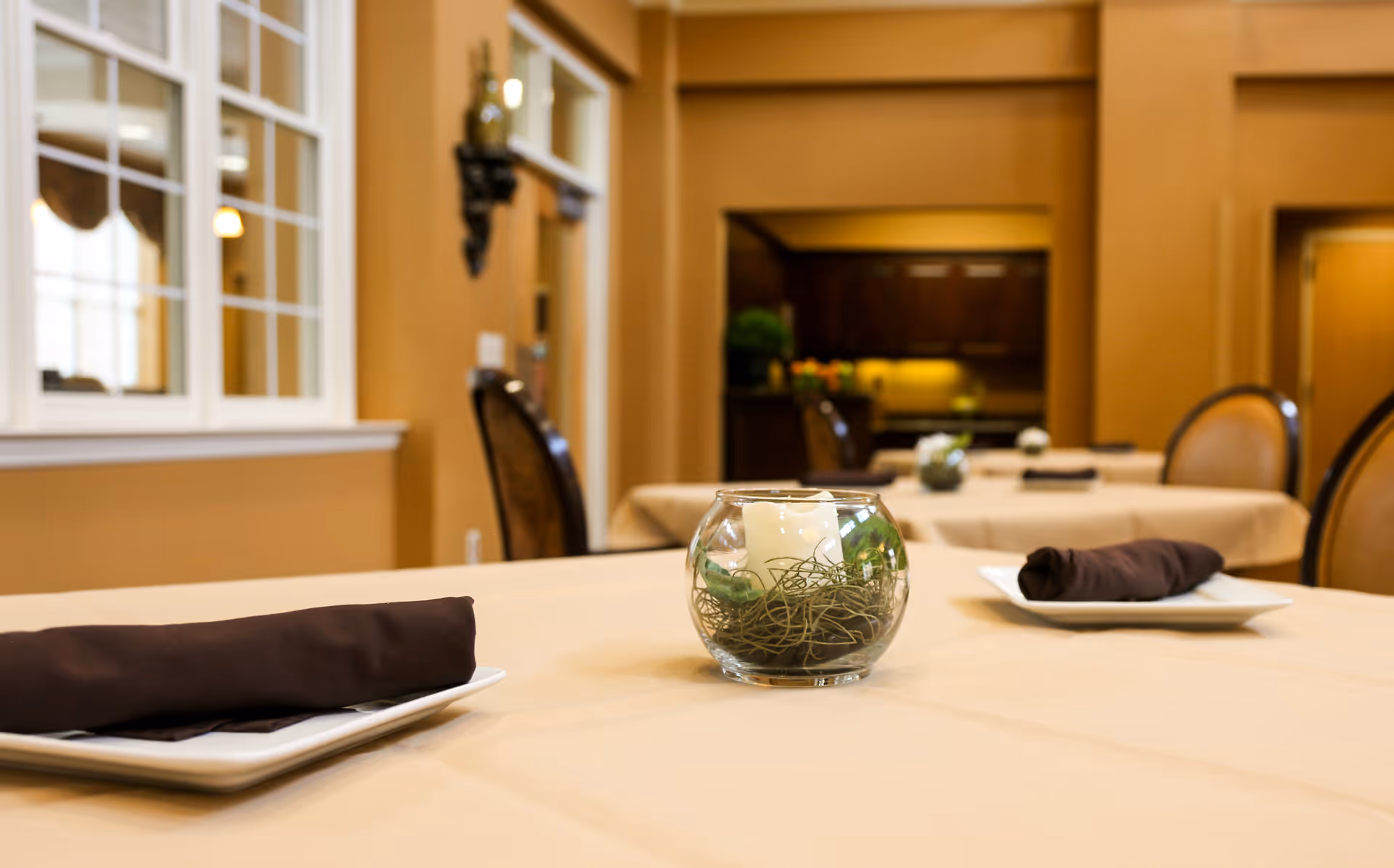 A dining area with tables covered in beige tablecloths, each set with a white plate and a neatly folded dark brown napkin. A small glass bowl centerpiece with a candle and decorative greenery is placed on the table in the foreground. The room has warm beige walls, a window with white trim, and chairs with cushioned backs.
