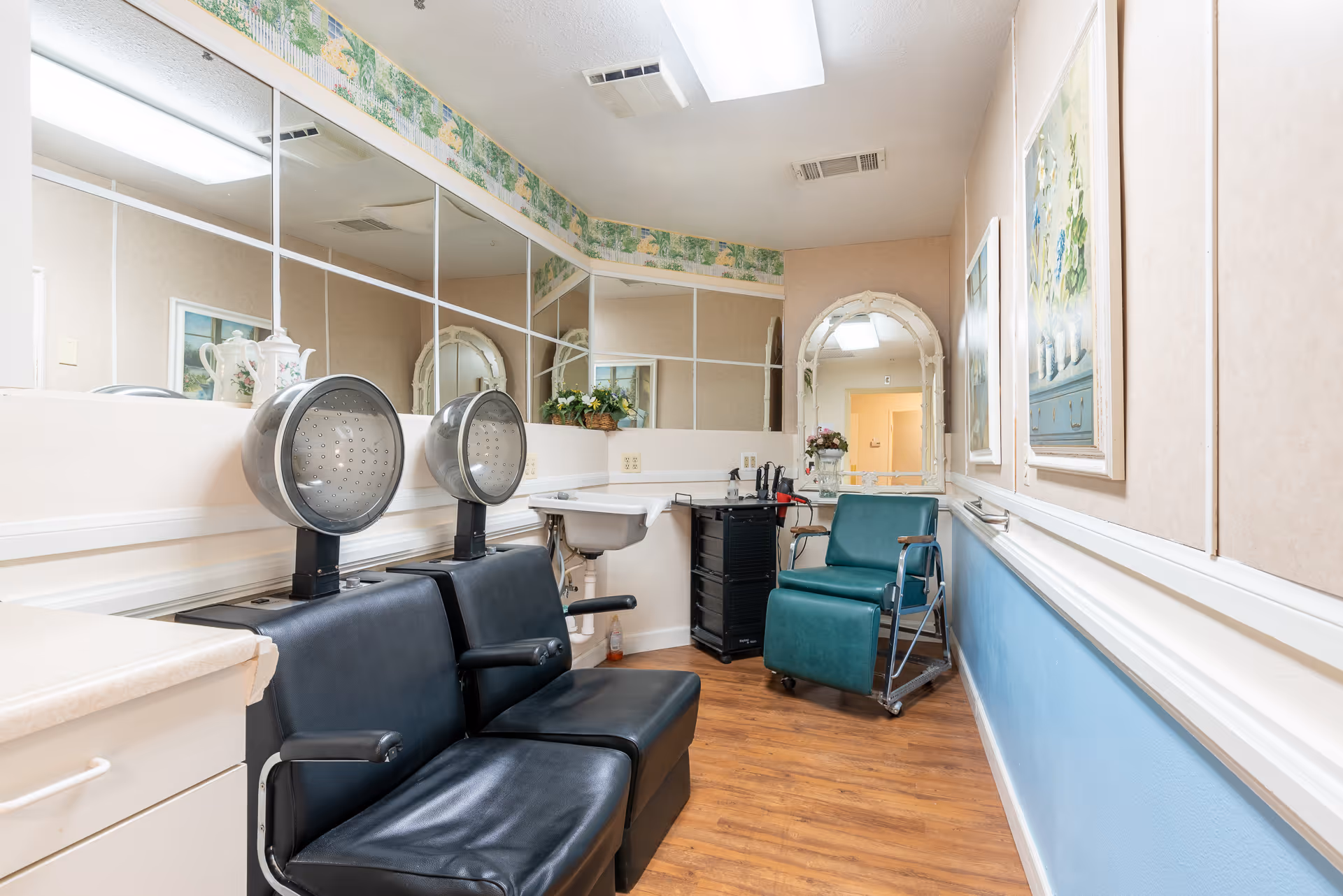 Interior view of a hair salon area in a senior living facility with two black salon chairs under hair dryers, a green salon chair on wheels, a small sink, a rolling cart with hair styling tools, large mirrors on the wall, floral wallpaper border near the ceiling, and framed artwork on the walls.