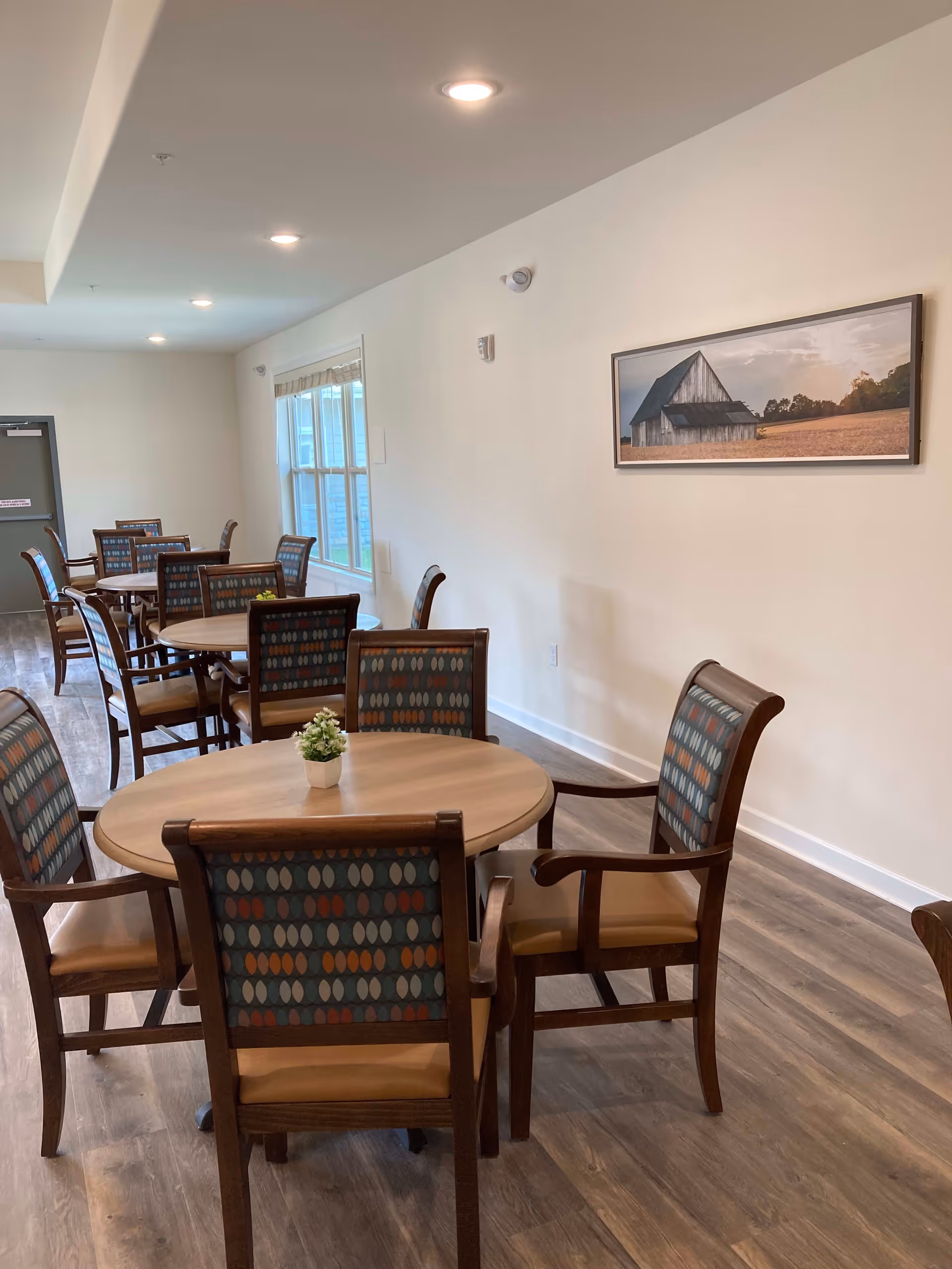 Interior view of a dining room with multiple round wooden tables and chairs with patterned upholstery. A small potted plant is placed on the nearest table. The room has wood flooring, white walls, recessed ceiling lights, a window with blinds, and a framed picture of a barn on the wall.