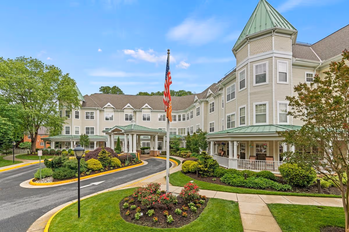 Exterior view of a senior living facility with beige siding and green roofs, featuring a circular driveway, landscaped gardens, an American flag, and a clear blue sky.