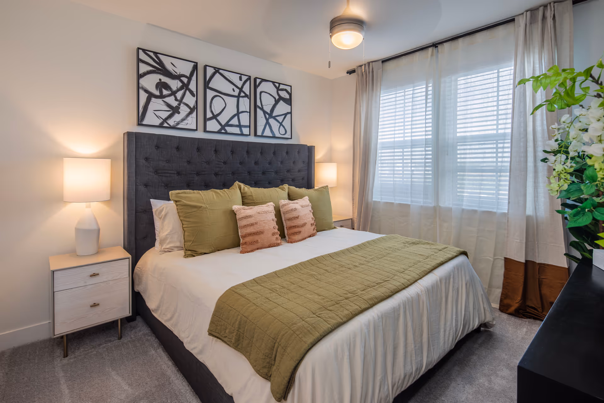 A neatly made bed with a dark gray tufted headboard, white sheets, green pillows, and a green quilted throw blanket in a bedroom. There are two bedside tables with white lamps on each side of the bed. Above the headboard are three abstract black and white framed artworks. A window with sheer white curtains and beige drapes is on the right side, and a ceiling fan with a light is visible above the bed.