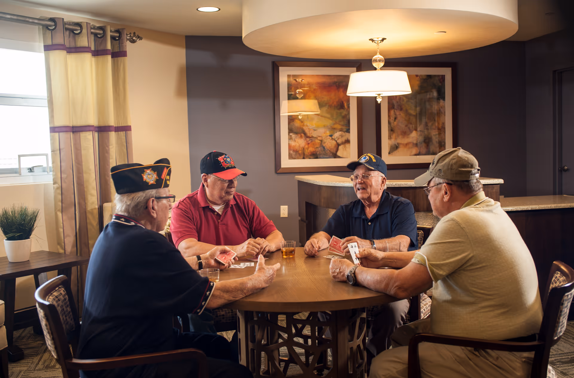 Four elderly men wearing hats sit around a round wooden table playing cards in a warmly lit room with framed artwork on the wall and a window with curtains.