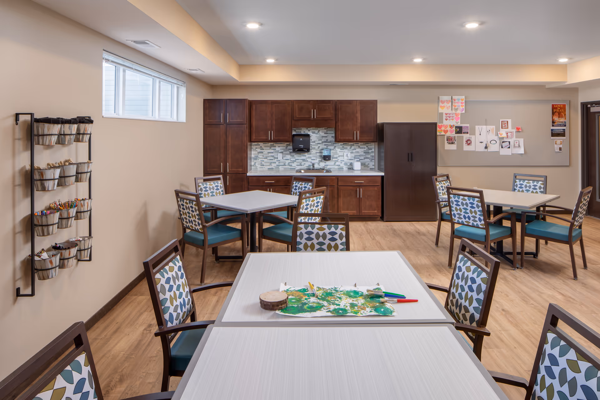 Communal activity/dining room with several tables and patterned chairs, a kitchenette with dark cabinets and a bulletin board on the far wall.