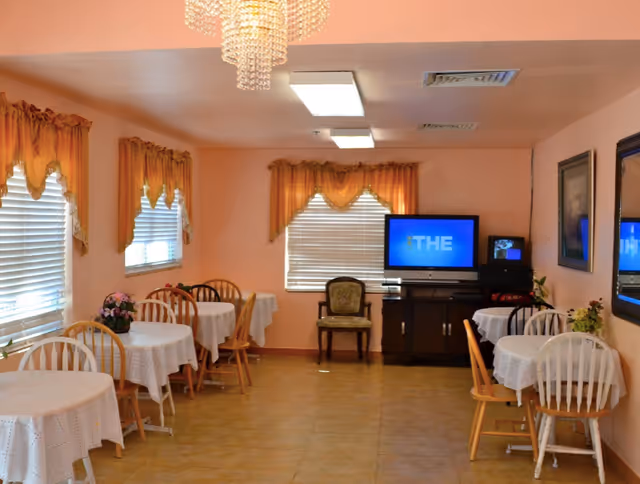 A small dining room with several round tables covered with white lace tablecloths, each surrounded by wooden chairs. The room has peach-colored walls, three windows with orange valances, a chandelier, and a television on a stand against the far wall. There are framed pictures on the walls and some floral decorations on the tables.