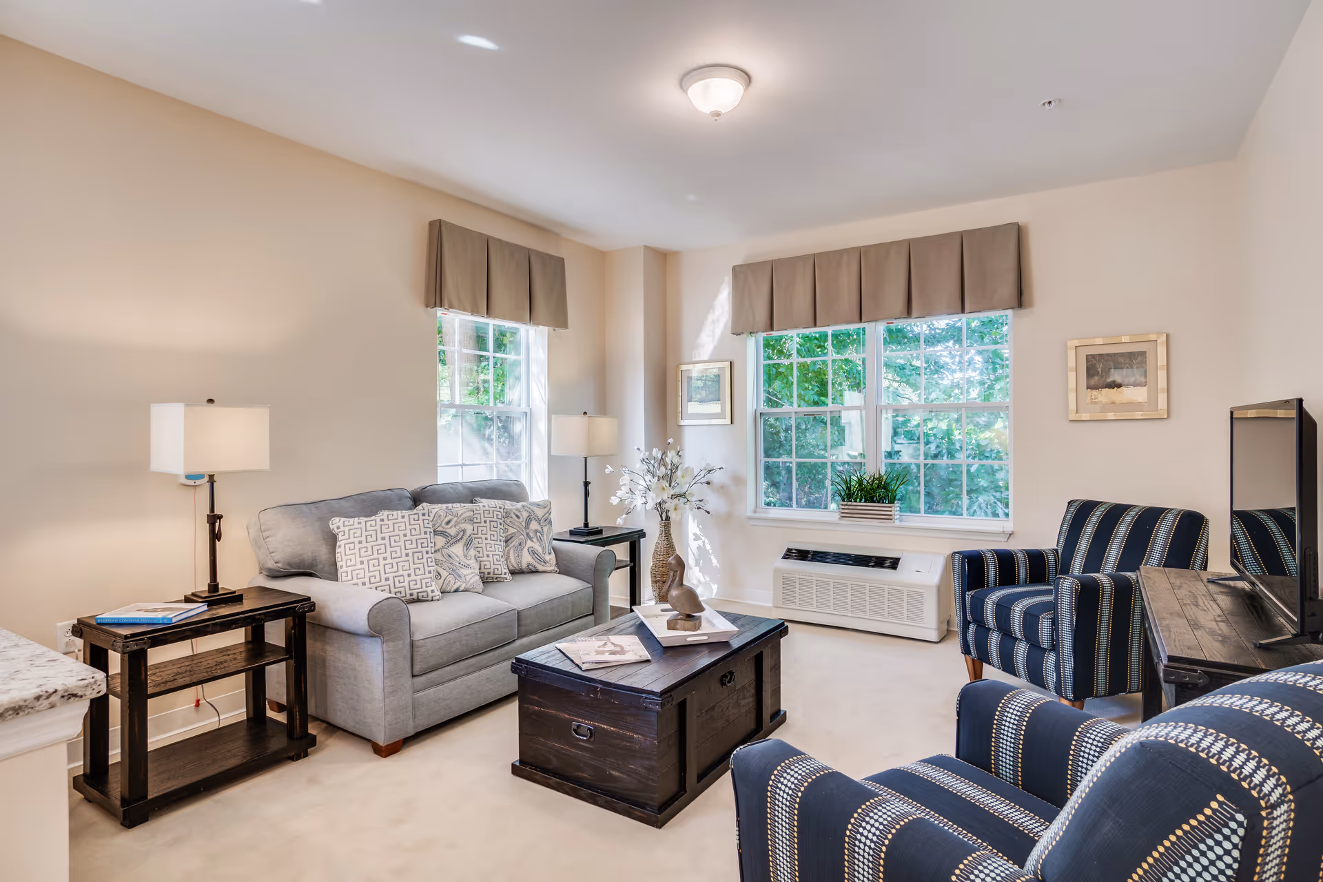 Well-lit living room with a gray sofa, two striped armchairs, a wooden coffee table, and large windows.