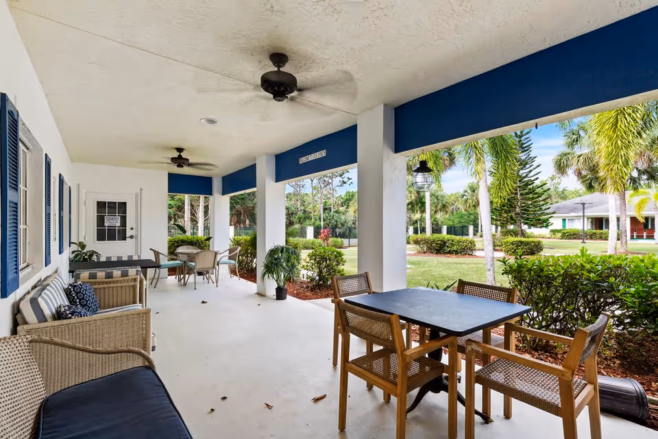 Covered outdoor patio area with ceiling fans, wicker seating with cushions, and tables with chairs. The patio overlooks a landscaped garden with palm trees and shrubs under a blue sky.