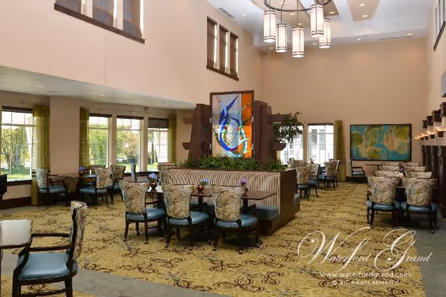 Spacious dining room with multiple tables and chairs arranged neatly. The room features large windows allowing natural light, decorative plants, and colorful artwork on the walls. A central seating area with a stained glass panel and pendant lights hanging from the ceiling add to the elegant ambiance.