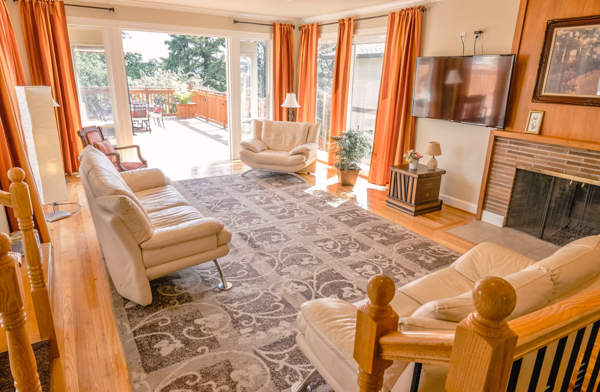 Bright living room with cream leather sofas, a patterned rug, fireplace, wall-mounted TV and sliding glass doors opening to a sunlit deck.