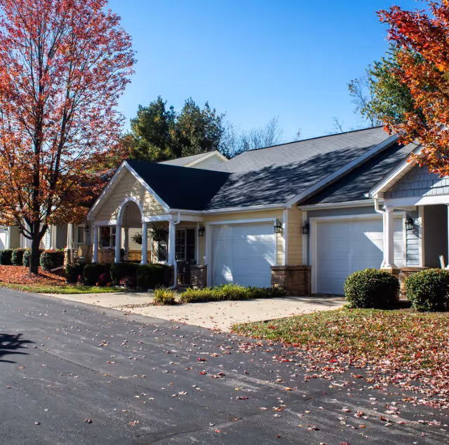 Exterior view of a senior living facility building with attached garages, surrounded by trees with autumn foliage and a clear blue sky.