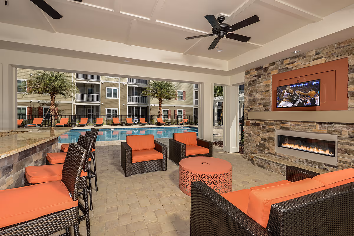 Covered outdoor seating area with wicker furniture featuring orange cushions, a round orange metal table, a stone fireplace with a mounted flat-screen TV showing a basketball game, and a ceiling fan. Beyond the seating area is a swimming pool surrounded by lounge chairs and palm trees, with a multi-story apartment building in the background.