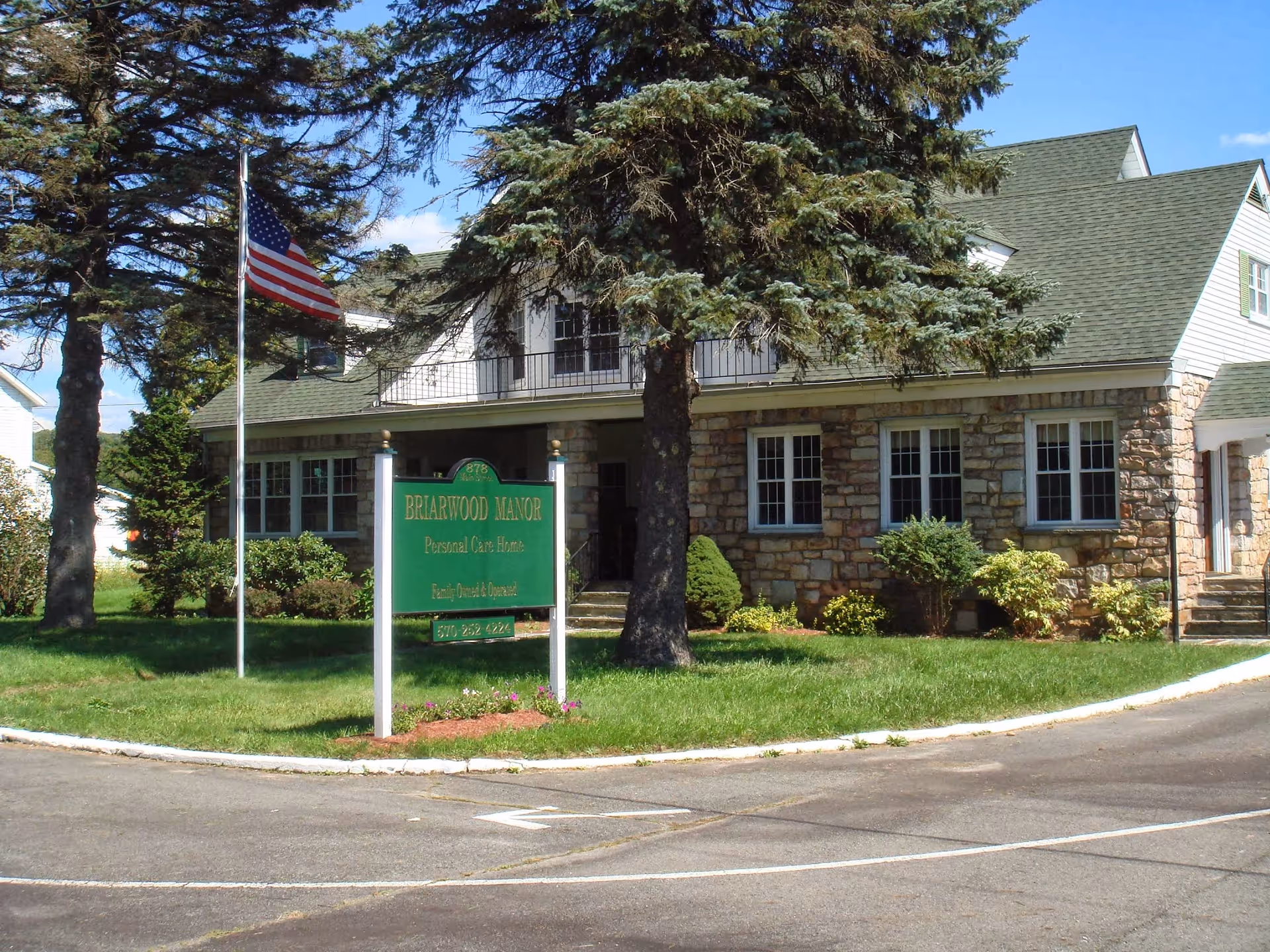 Exterior view of a stone and siding building with a green roof, surrounded by trees and shrubs. A green sign in front reads 'Briarwood Manor Personal Care Home' with additional text below. An American flag is flying on a flagpole near the sign.