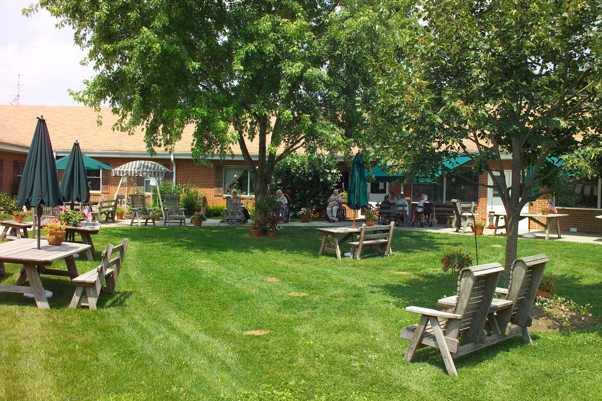 Outdoor garden area of Hickory House Nursing Home with green grass, several wooden picnic tables with umbrellas, wooden benches, and mature trees providing shade. Several elderly people are sitting and relaxing near the building in the background.
