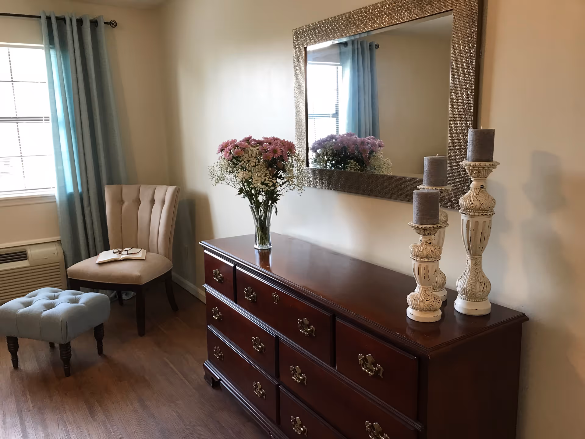 A cozy interior corner featuring a wooden dresser with brass handles, decorated with a vase of pink and white flowers and three ornate white candle holders with gray candles. Above the dresser is a large rectangular mirror reflecting part of the room. To the left, there is a beige upholstered chair with a book and glasses resting on it, a small blue tufted ottoman, a window with blue curtains, and a wall-mounted air conditioning unit below the window.