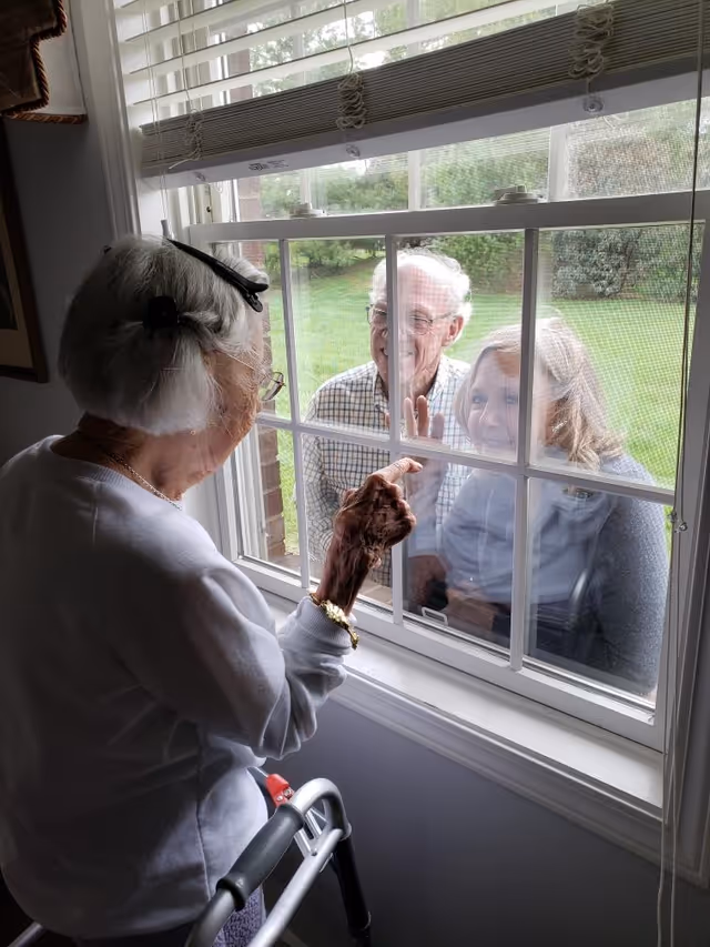 An elderly woman inside a room uses a walker and touches a window screen while looking outside. Outside the window, an elderly man and woman are smiling and looking in, with the woman in a wheelchair.