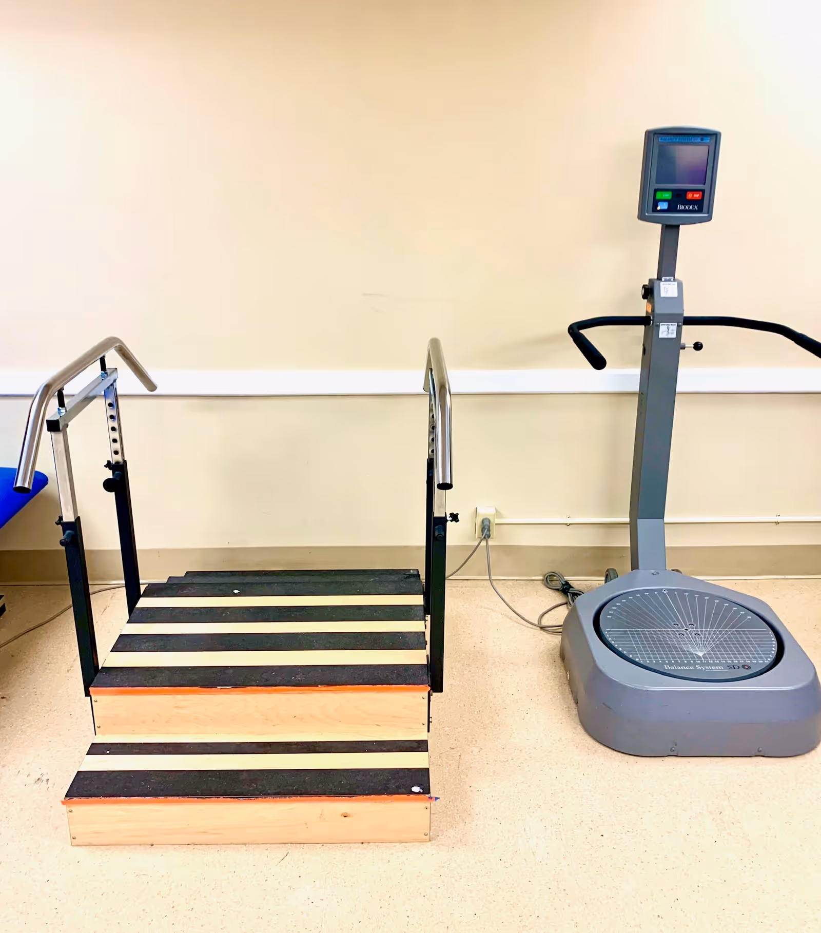 Physical therapy equipment including a short wooden practice staircase with handrails and a balance/vibration platform against a beige wall.