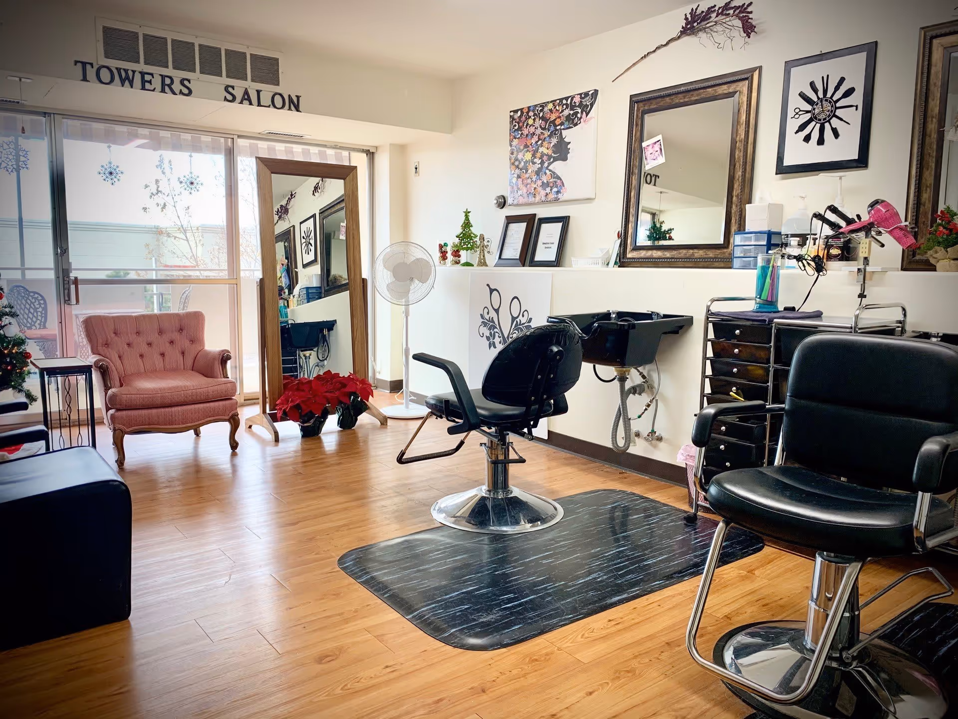 Interior of a salon room with wooden flooring, two black salon chairs, a black hair washing sink, a large standing mirror, a pink upholstered armchair, a small table, and various wall decorations including framed pictures and a sign that reads 'TOWERS SALON'. There are also holiday decorations like a small Christmas tree and poinsettia plants.