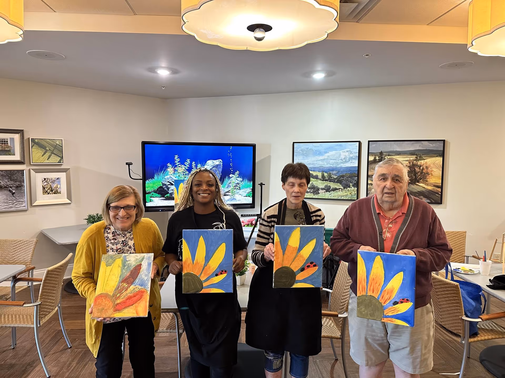Four adults standing indoors in a well-lit room holding up paintings of sunflowers with a ladybug on the petals. Behind them are framed landscape paintings and a TV screen displaying an underwater scene. The room has tables and chairs arranged around them.