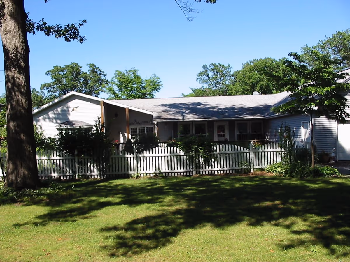 Single-story house with light-colored siding, a gray roof, and a white picket fence surrounding a garden area with bushes and trees, under a clear blue sky.