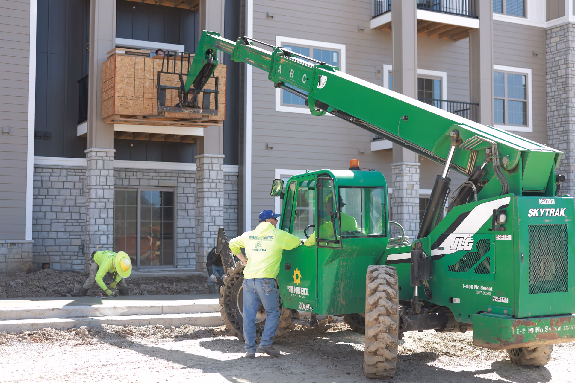 Construction workers operating a green SkyTrak forklift and working on the ground outside a senior living facility building with stone and siding exterior walls and balconies.