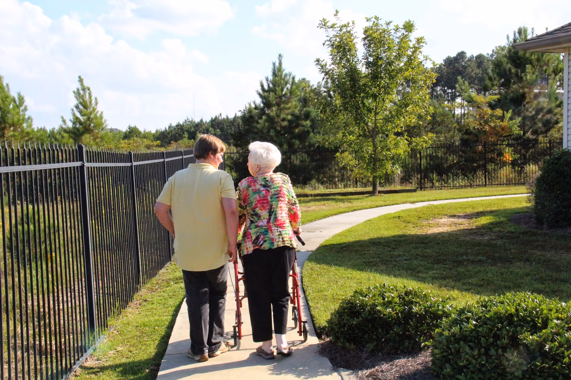 An elderly woman using a walker is walking on a paved path outdoors accompanied by a younger person. They are walking away from the camera along a fenced pathway surrounded by grass, bushes, and trees under a partly cloudy sky.