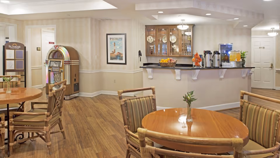 Communal dining area with round wooden tables and bamboo-style chairs, a service counter with beverages, and a vintage jukebox.