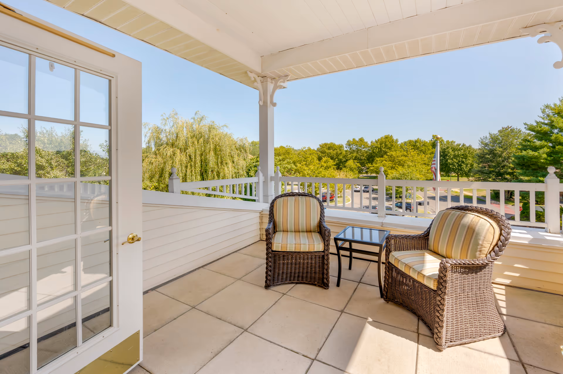 A covered outdoor balcony with two wicker chairs featuring striped cushions and a small glass-top table between them. The balcony has white railings and overlooks a parking lot and green trees under a clear blue sky.