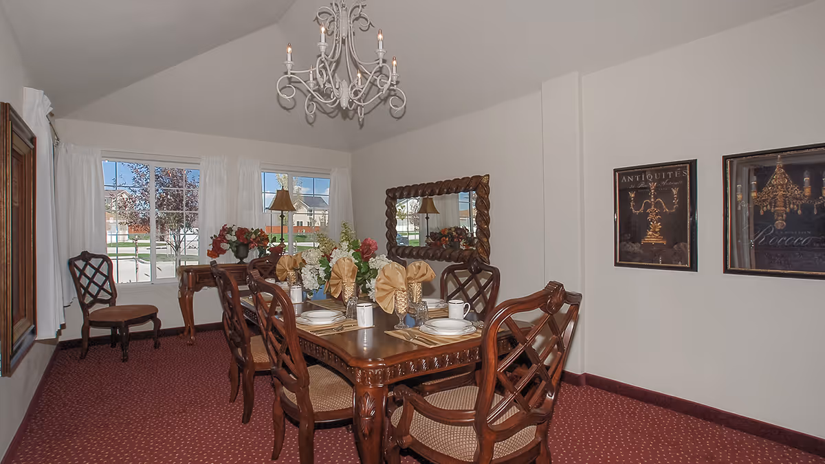 A formal dining room with a wooden dining table set for six people. The table is decorated with floral centerpieces and folded napkins. There are six wooden chairs with cushioned seats around the table. The room has a red carpet with a small pattern, white walls, and a large mirror on one wall. Two framed pictures hang on another wall. A chandelier with lit candles hangs from the ceiling. Large windows with white curtains let in natural light and show a view of houses and trees outside.