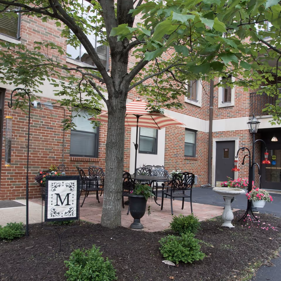 Outdoor patio area at Mount Carmel Communities featuring a large tree, black metal chairs and tables with a red and white striped umbrella, flower pots, a birdbath, and a decorative sign with the letter M. The background shows a brick building with windows and a door.
